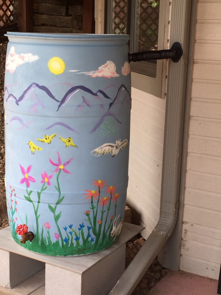 A 55-gallon barrel painted with flowers and mountains sits on a stand and is connected to the rain gutter of a home.